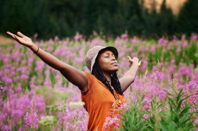 A woman with her eyes closed and arms outstretched stands in a field of purple wildflowers with a lush, vibrant green forest in the background.