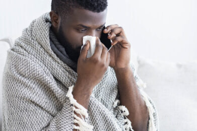 A man wrapped in a blanket speaks on his phone while holding a tissue to his nose, illustrating a remote medical consultation from home.