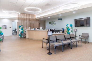 The modern reception area of Apple Valley Health Center, featuring a light wood front desk, grey seating, and celebratory green, blue, and white balloon pillars.